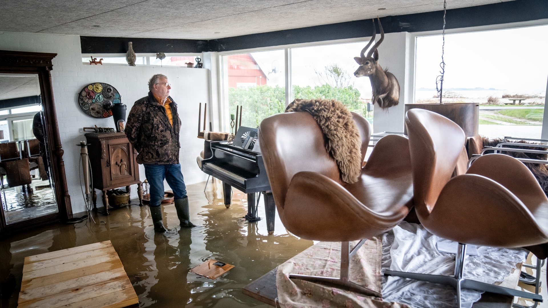Mand i gummistøvler og overtøj står ved et flygel i en stue, hvor gulvet er oversvømmet af vand fra stormflod.